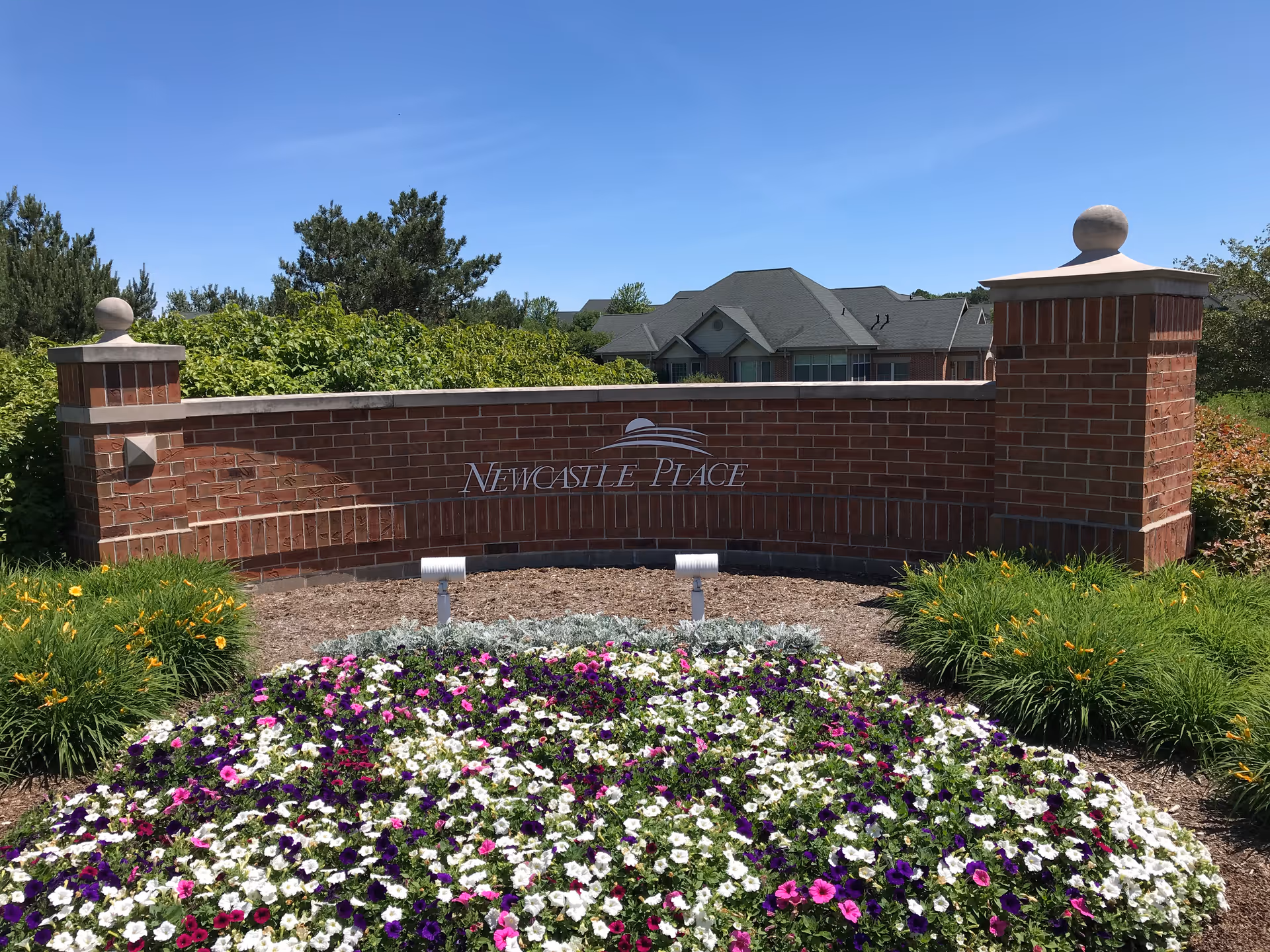 Brick entrance sign reading Newcastle Place with flowers, shrubs, and a building in the background.