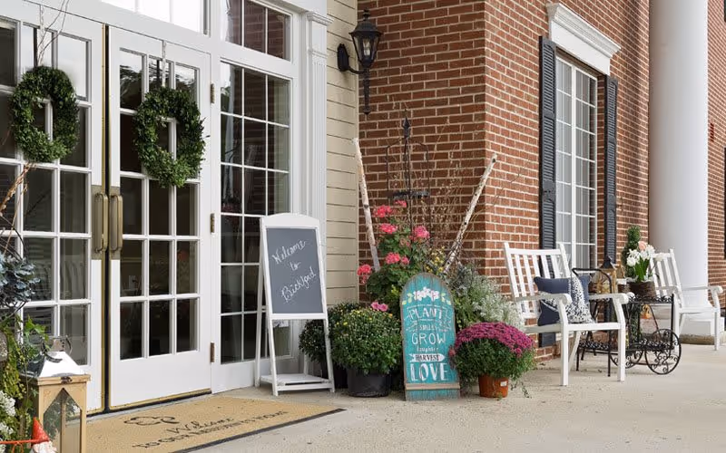 Entrance of a senior living facility with double glass doors adorned with green wreaths. There is a welcome mat on the ground, a small chalkboard sign that says 'Welcome to RoseBridge', and various potted plants and flowers arranged near the doorway. White chairs and a small table with decorative items are placed along the brick wall under white columns.