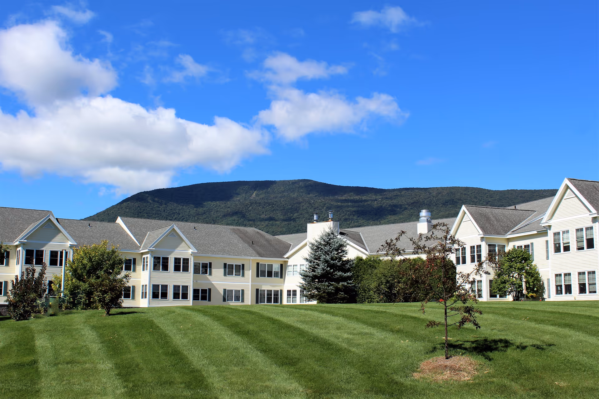 Exterior view of a large, two-story senior living facility with beige siding and multiple windows, set against a backdrop of green hills and a blue sky with scattered clouds. The foreground features a well-maintained lawn with striped mowing patterns and several small trees and shrubs.