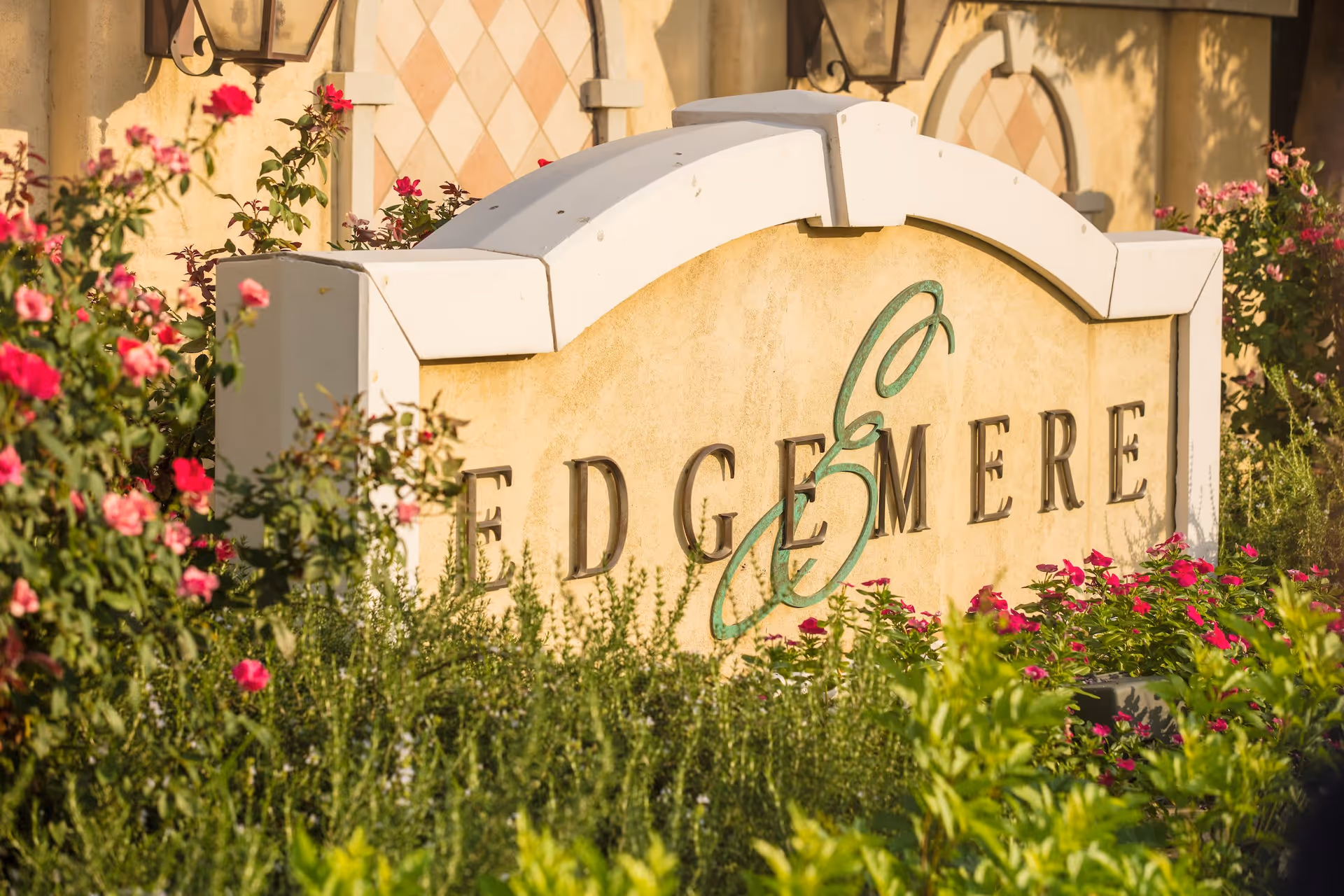 A decorative stone sign with the word 'EDGEMERE' partially visible, surrounded by lush green plants and blooming pink flowers, with a building wall featuring diamond-patterned tiles in the background.