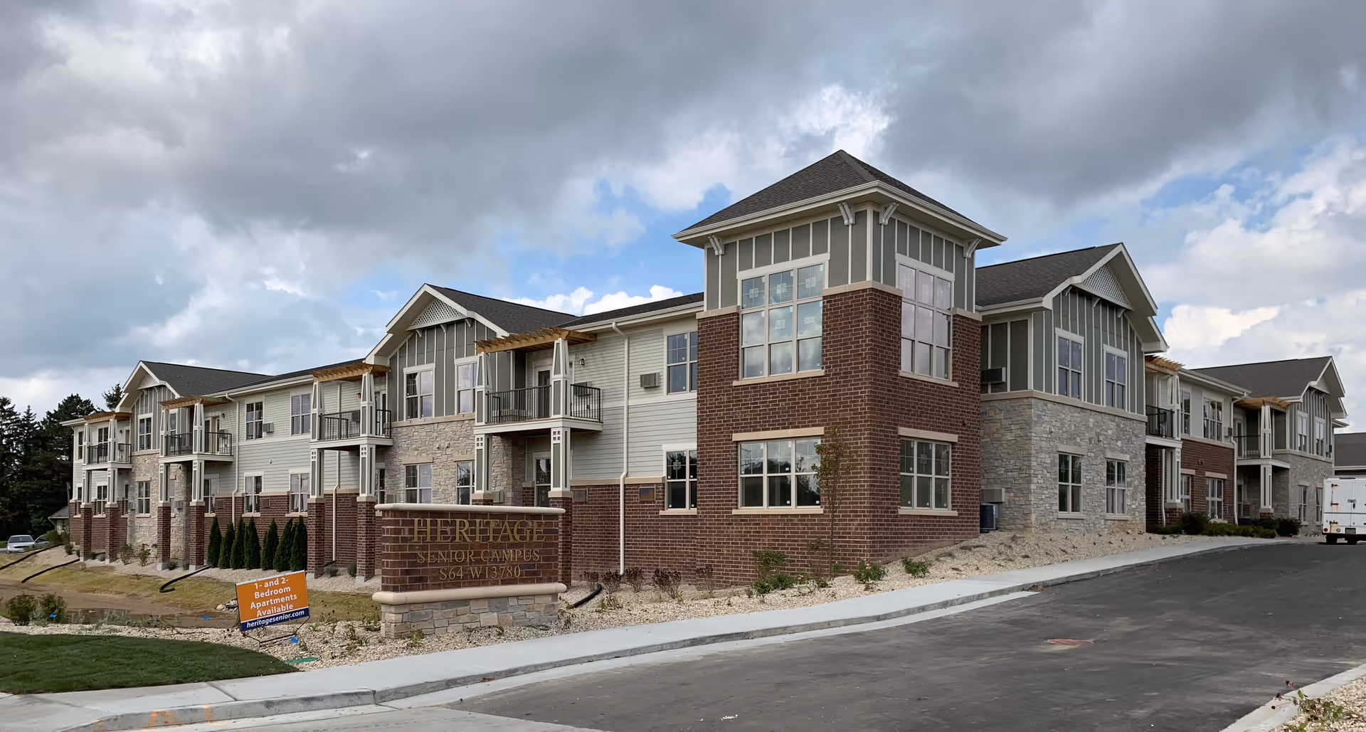 Exterior view of a large, modern senior living facility building with multiple windows and balconies. The building features a combination of brick, stone, and siding materials with a paved driveway in front. A sign near the entrance reads 'Heritage Senior Campus' along with an address and a smaller sign advertising 1 and 2 bedroom apartments available.