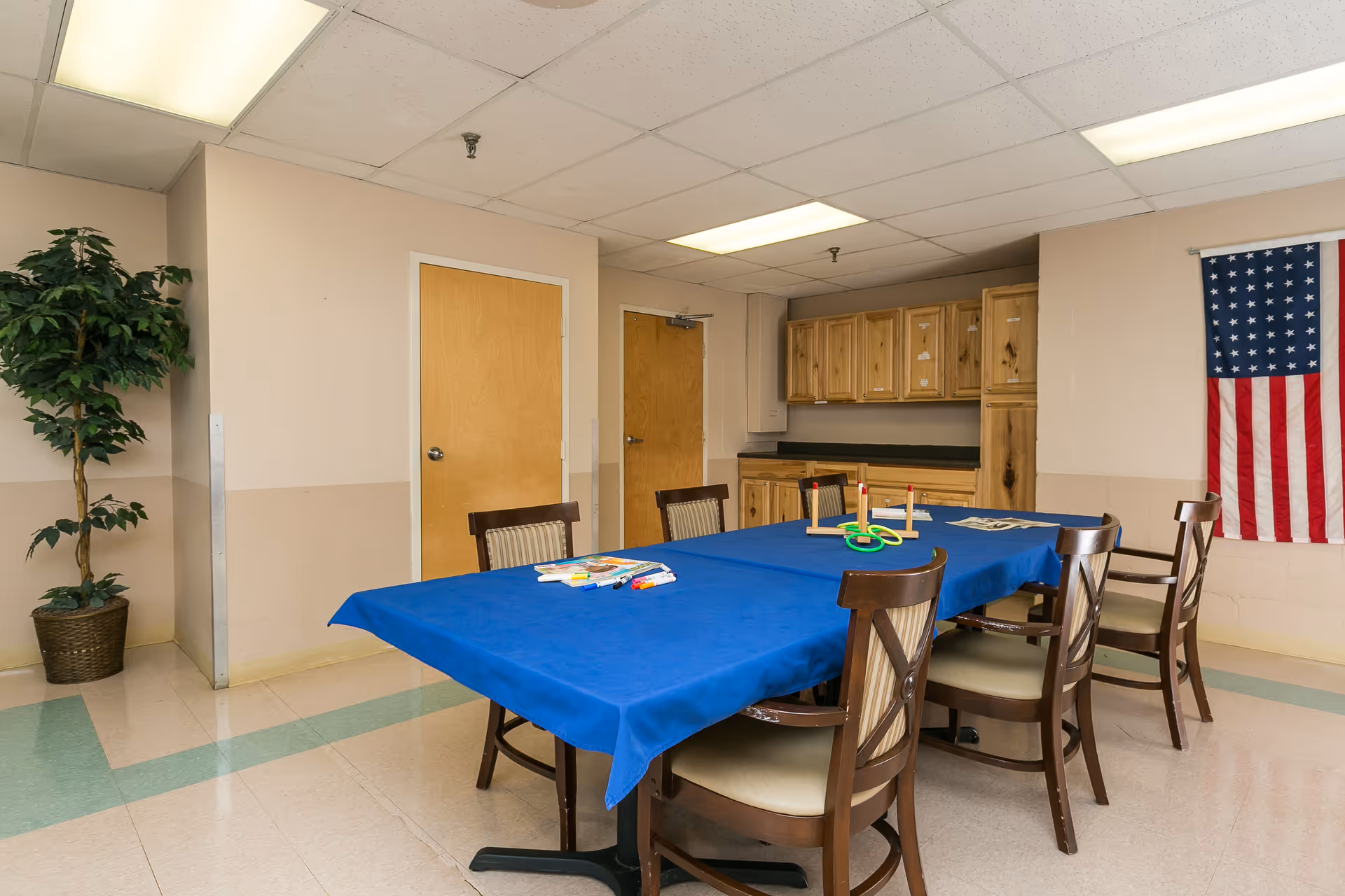 A room with a rectangular table covered with a blue tablecloth surrounded by six wooden chairs with cushioned seats. On the table are some markers, papers, and a ring toss game. The room has beige walls, two wooden doors, wooden cabinets with a countertop, a potted plant in the corner, and an American flag hanging on the wall.