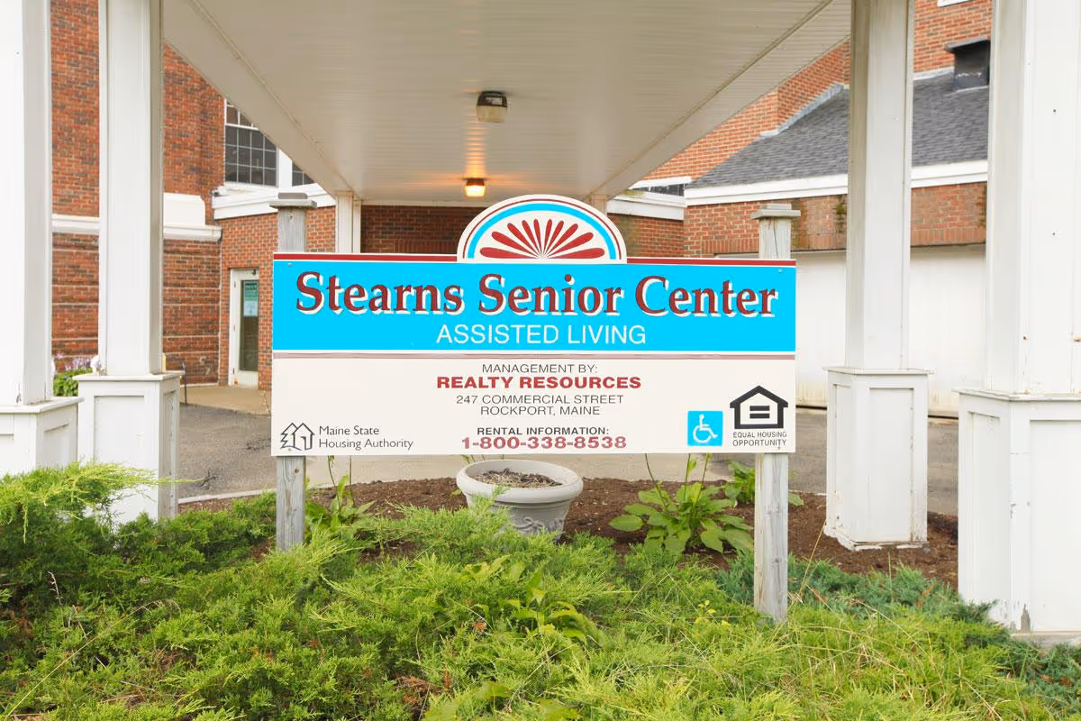 A sign for Stearns Senior Center Assisted Living located under a covered walkway with white pillars. The sign includes management information for Realty Resources in Rockport, Maine, rental information phone number, and logos for Maine State Housing Authority and Equal Housing Opportunity. There are green bushes and plants in front of the sign.