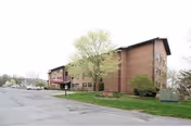 Brick senior living building front with parking and trees.