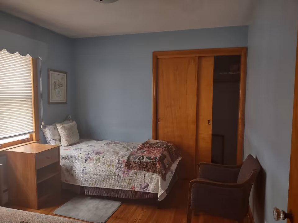 Cozy blue bedroom with bed, window blinds, dresser, closet doors, and two chairs.