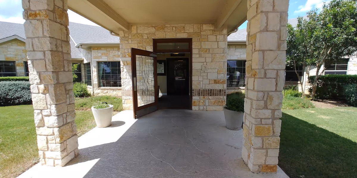 Stone-covered entrance with two pillars and an open decorative glass door leading into the building, flanked by planters and lawn.