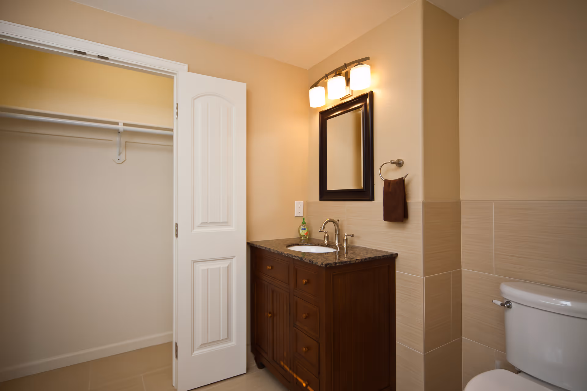 A bathroom with a wooden vanity featuring a granite countertop and a sink. Above the sink is a rectangular mirror with a dark frame and a three-light fixture. To the right of the vanity is a white toilet. On the left side, there is an open door revealing an empty closet with a hanging rod. The walls are painted beige and partially tiled.