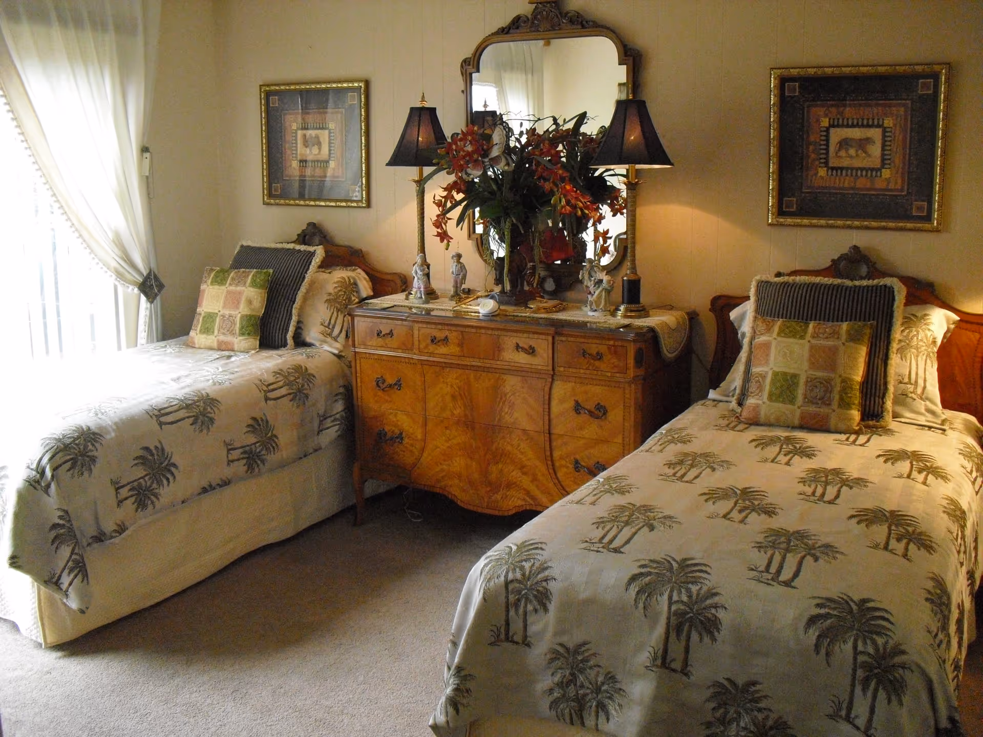 Two twin beds with palm-print bedspreads flank a wooden dresser topped with a mirror, lamps, and a floral arrangement.