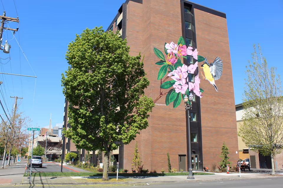 Brick apartment building with a large pink-flower-and-bird mural, a tree in front, and a clear blue sky.
