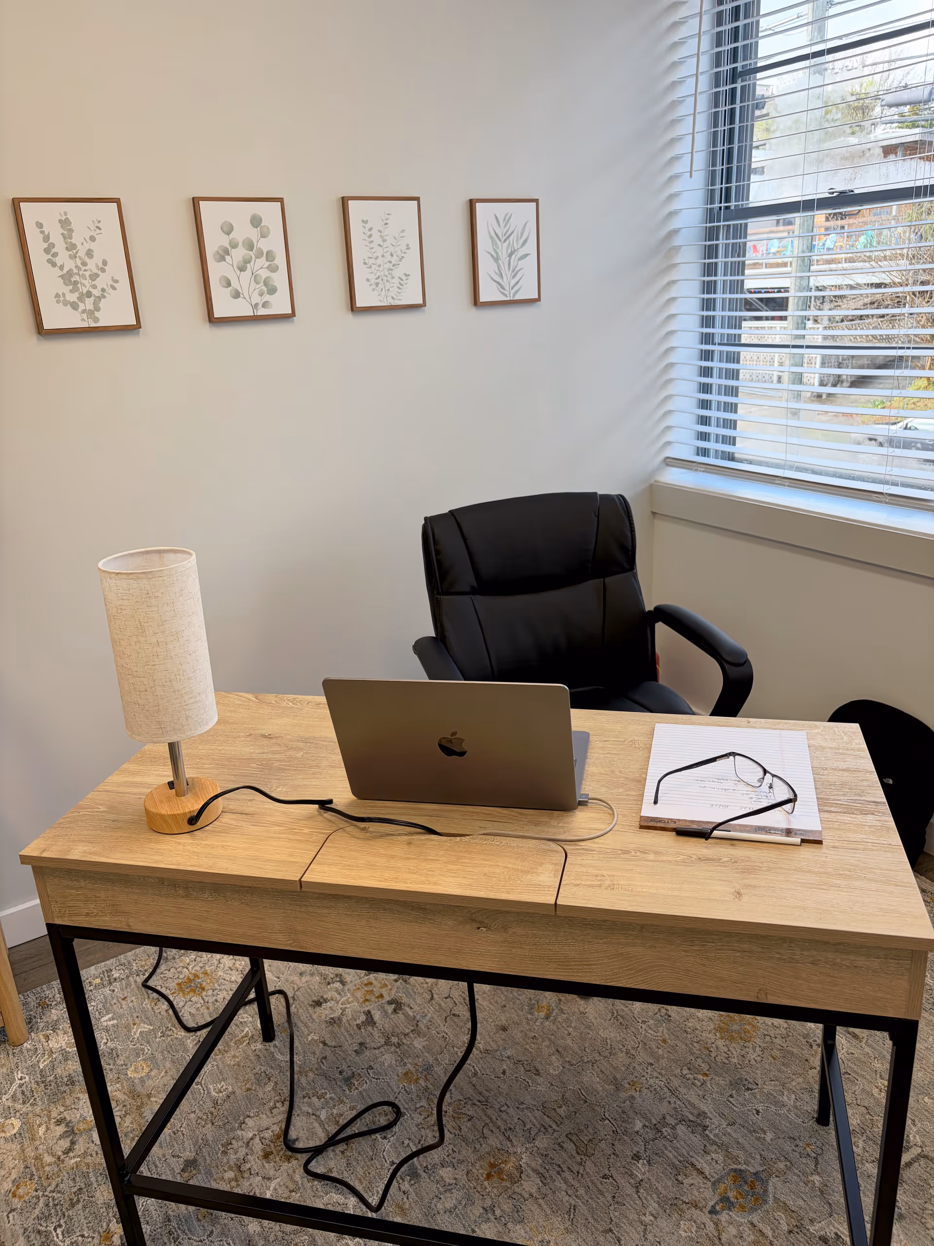 A light wooden office desk with a laptop, lamp, notebook, and glasses by a window.