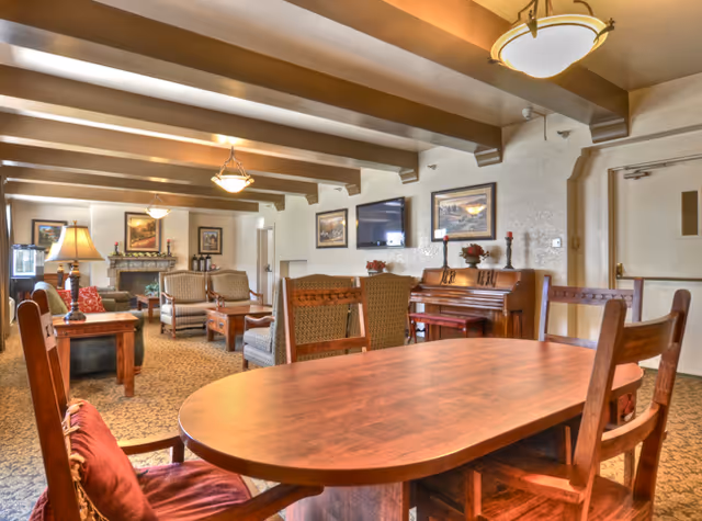 A cozy living room area in a senior living facility featuring a wooden dining table with chairs in the foreground, upholstered armchairs and sofas arranged around a fireplace, framed artwork on the walls, a piano against one wall, and warm lighting from ceiling fixtures and table lamps.