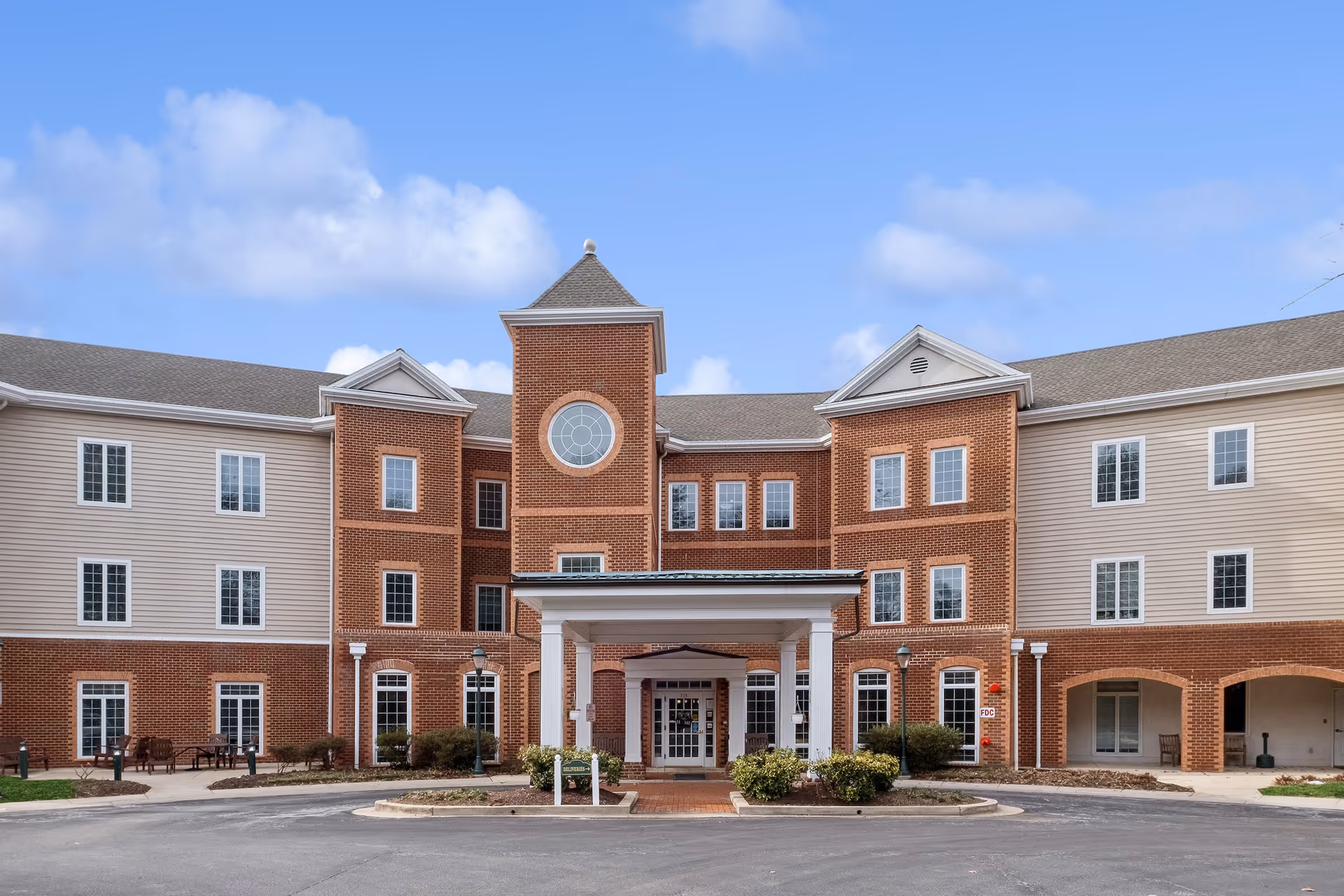 Front exterior view of HeartLands Assisted Living at Severna Park, a three-story building with a combination of red brick and beige siding, featuring a central entrance with a covered portico and a circular window above it under a peaked roof. The sky is clear with a few clouds.