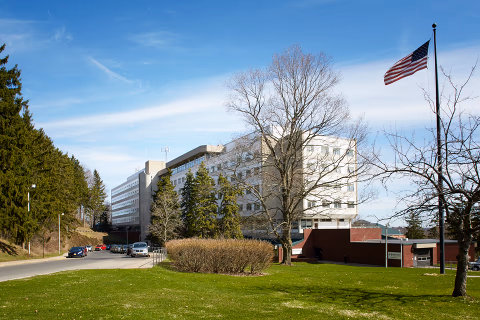 Exterior view of a senior living center with flagpole, trees, and a parking lot.