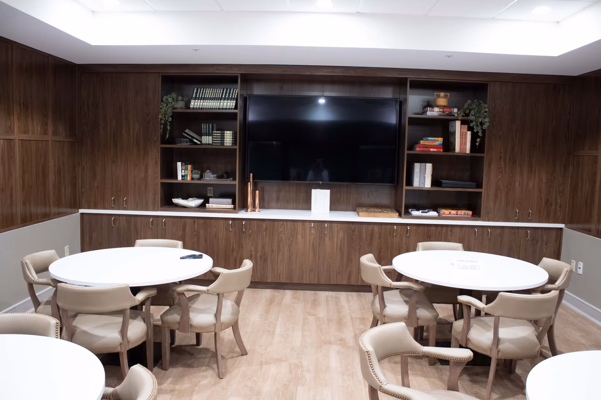 Communal dining/activity room with round white tables, beige upholstered chairs, and a large TV set into wood built-in cabinets.