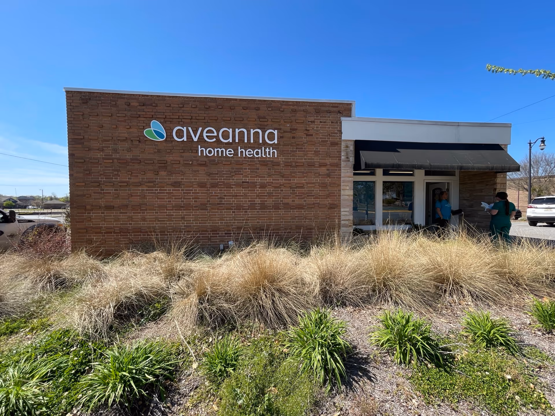 Brick building with Aveanna Home Health sign and entrance, two people near doorway.