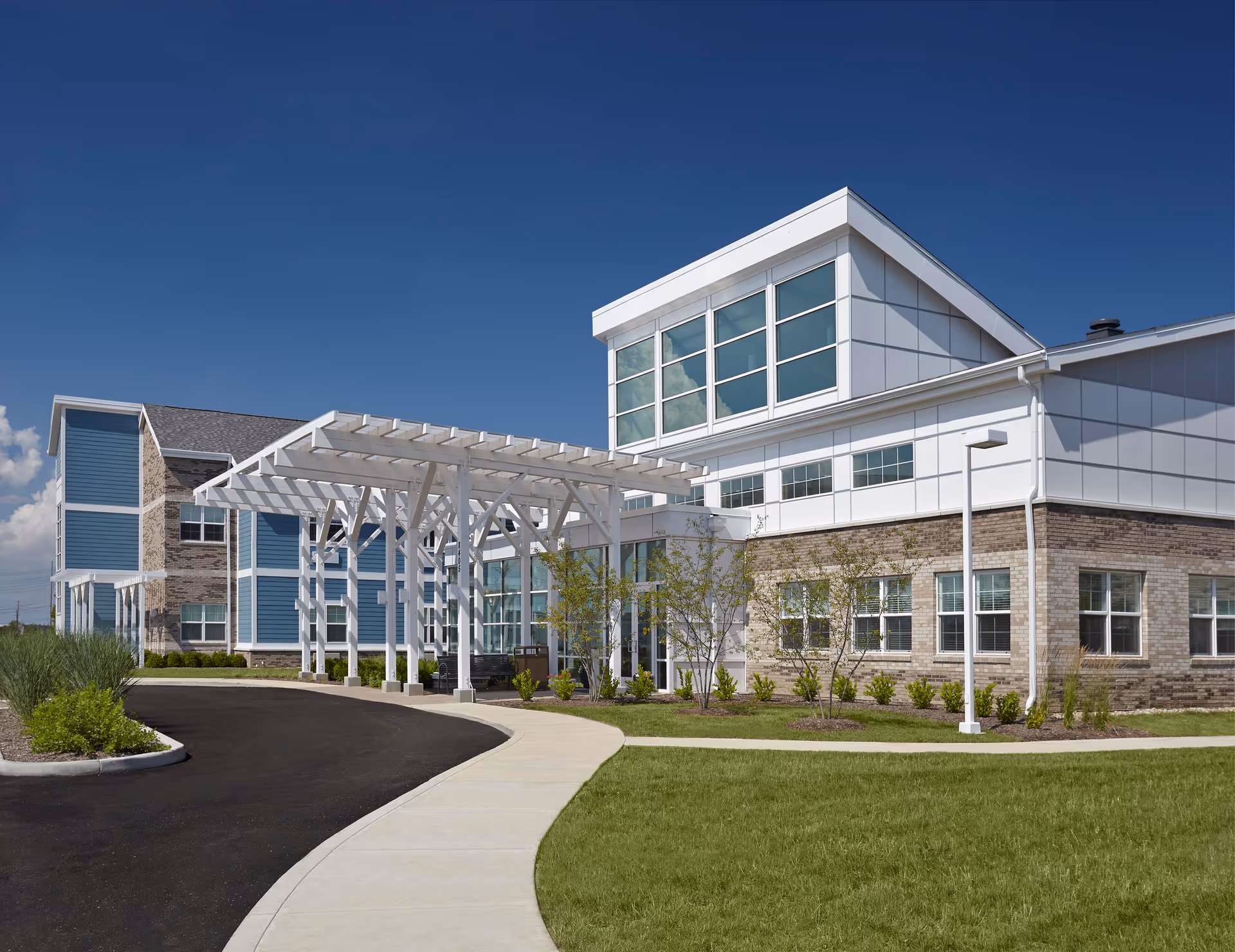 Front entrance of a modern senior living facility with a white covered porte-cochere, blue siding, brick accents and large windows under a clear blue sky.