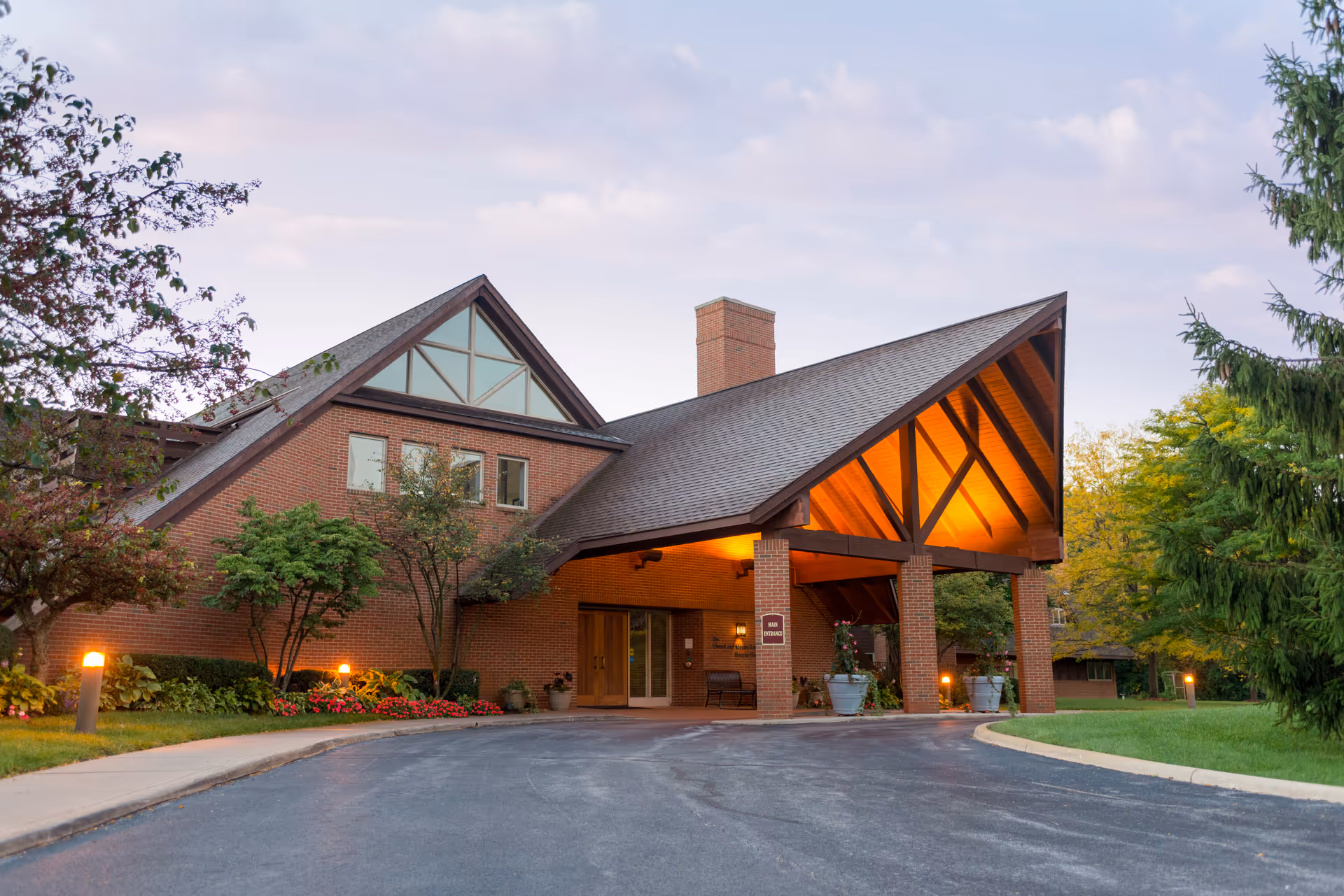Brick building with large triangular roof entrance and landscaped front, lit warmly at dusk.