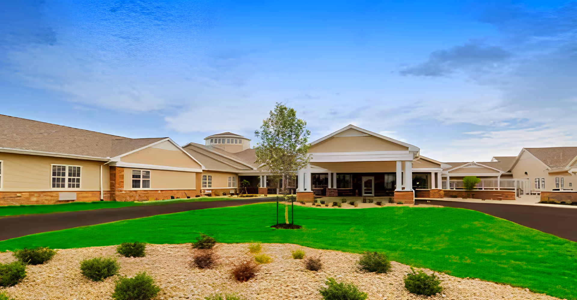 Front exterior of a single-story senior living facility with a covered entrance, green lawn, and landscaped beds under a blue sky.