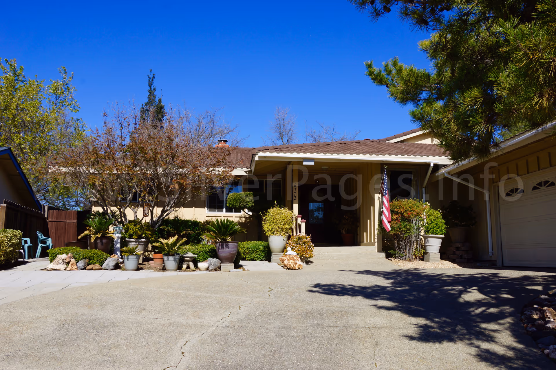 Single-story beige senior living home facade with potted plants and an American flag.