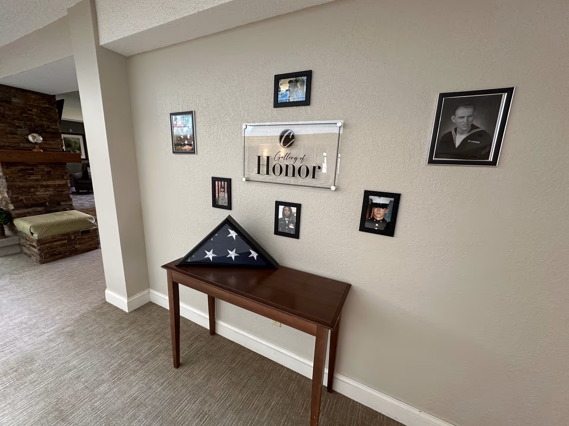 A hallway corner in a senior living facility featuring a wooden table with a folded American flag in a triangular display case. Above the table is a sign that reads 'Gallery of Honor' surrounded by framed photographs of military personnel on a beige textured wall. To the left, part of a stone fireplace and seating area is visible.