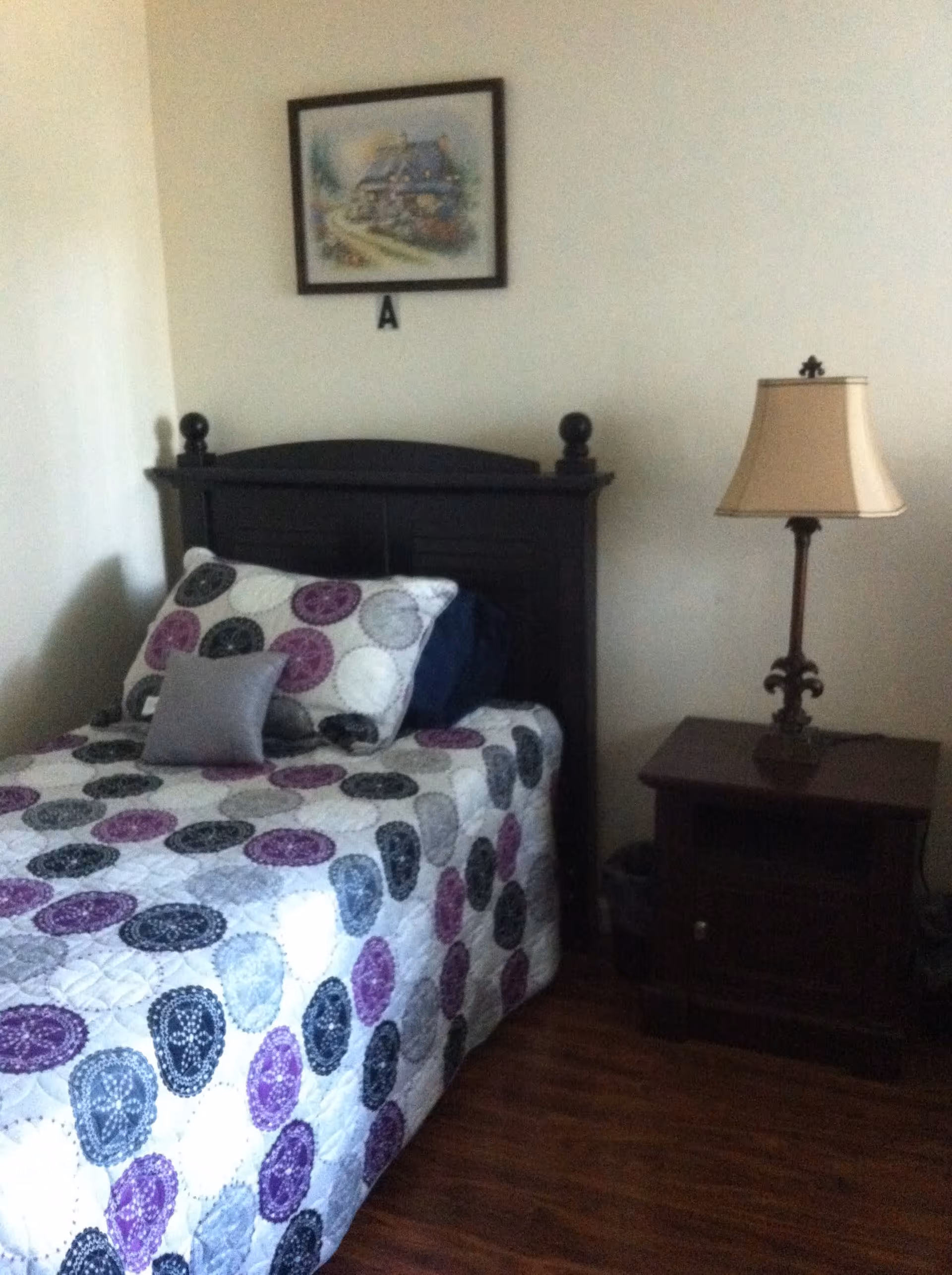 A small bedroom with a single bed featuring a quilted bedspread with circular patterns in purple, black, and gray. The bed has a dark wooden headboard and is accompanied by a matching nightstand with a decorative lamp. A framed picture hangs on the wall above the bed.