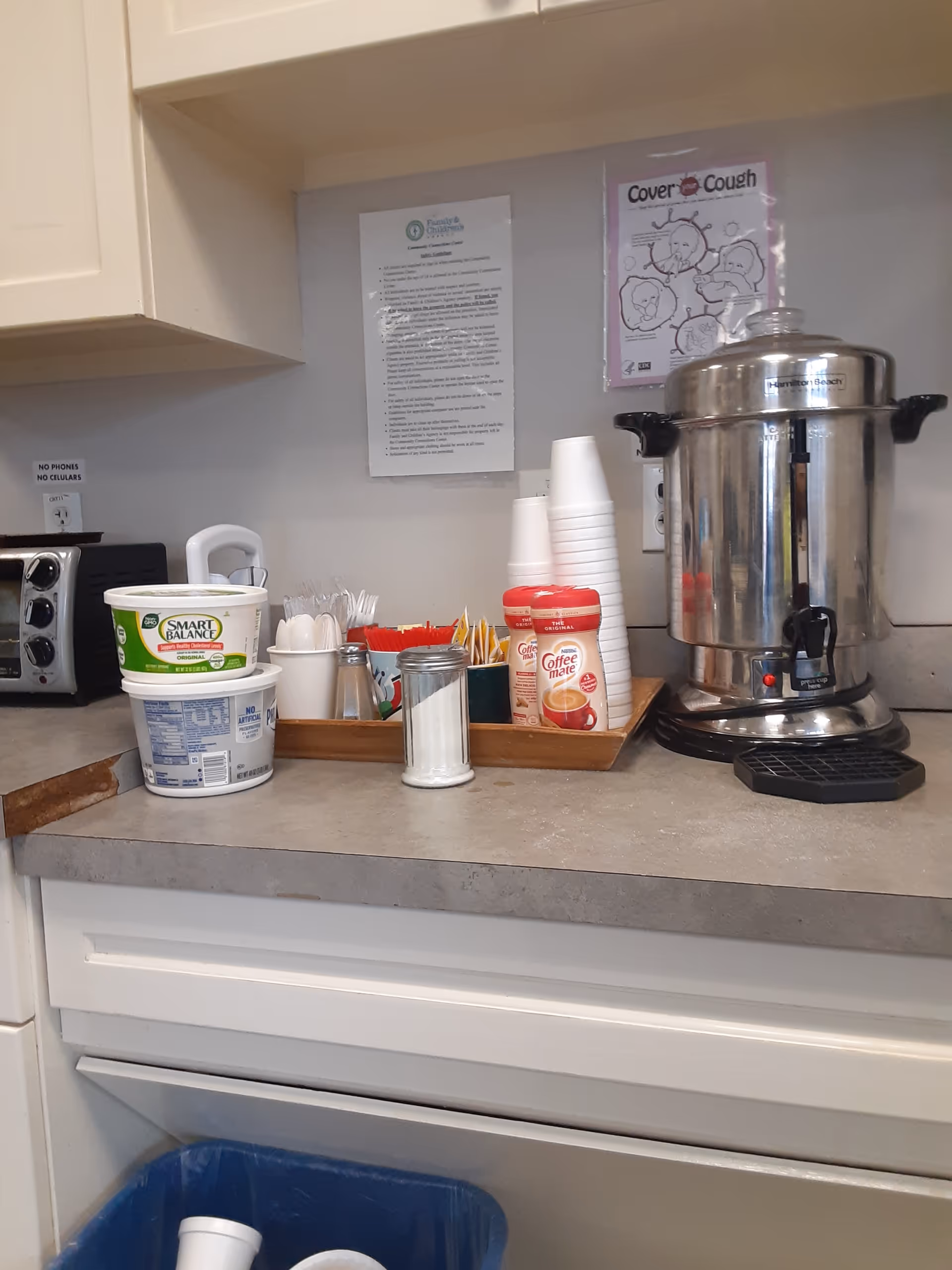 A kitchen counter with a large coffee urn, cups, and condiments.