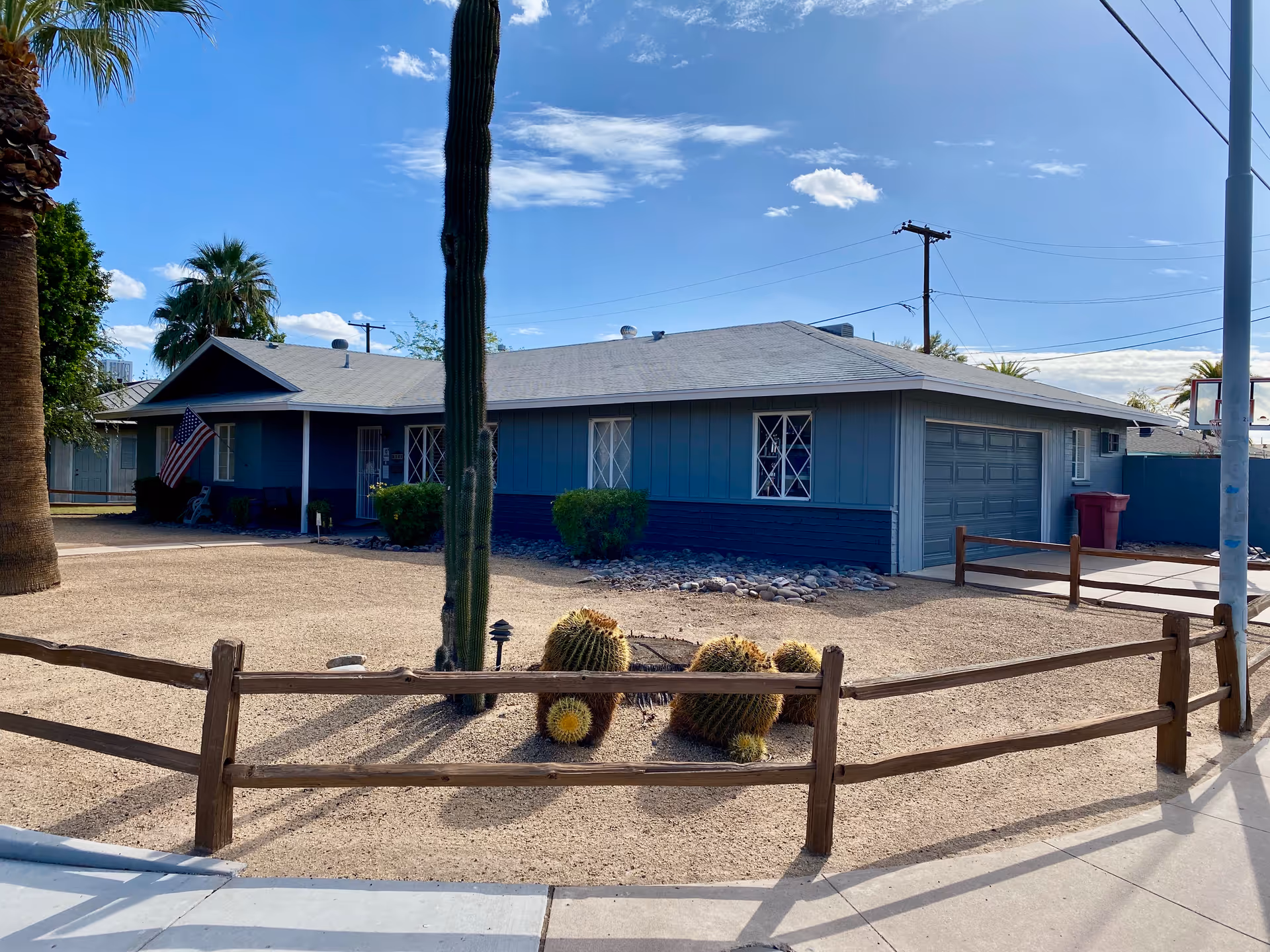 Single-story blue house with a gray roof surrounded by a gravel yard with cacti and a wooden fence. There is an American flag near the entrance and a garage on the right side. The sky is clear with a few clouds.