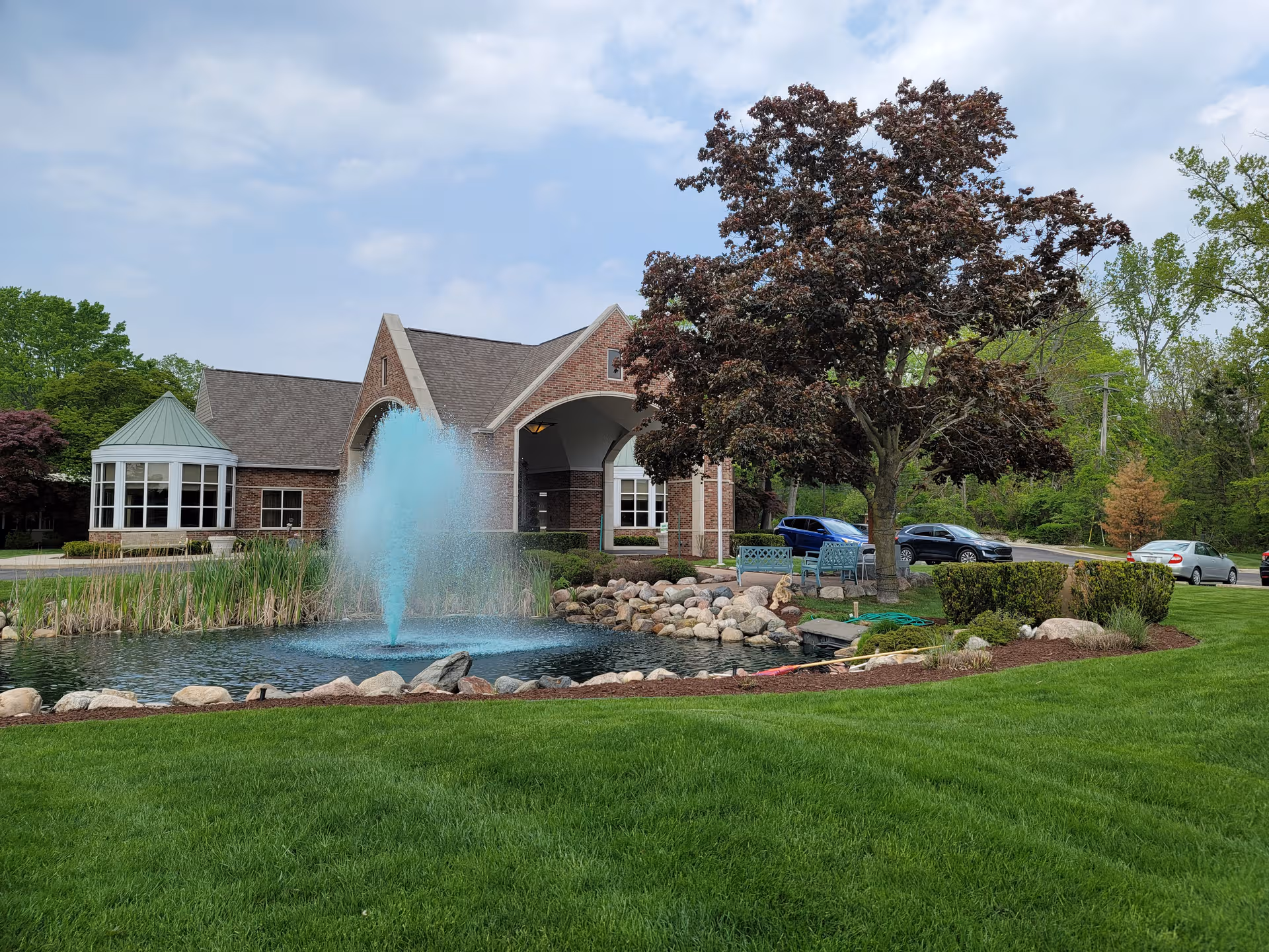 Brick senior living building with a central fountain pond, manicured lawn, and trees.