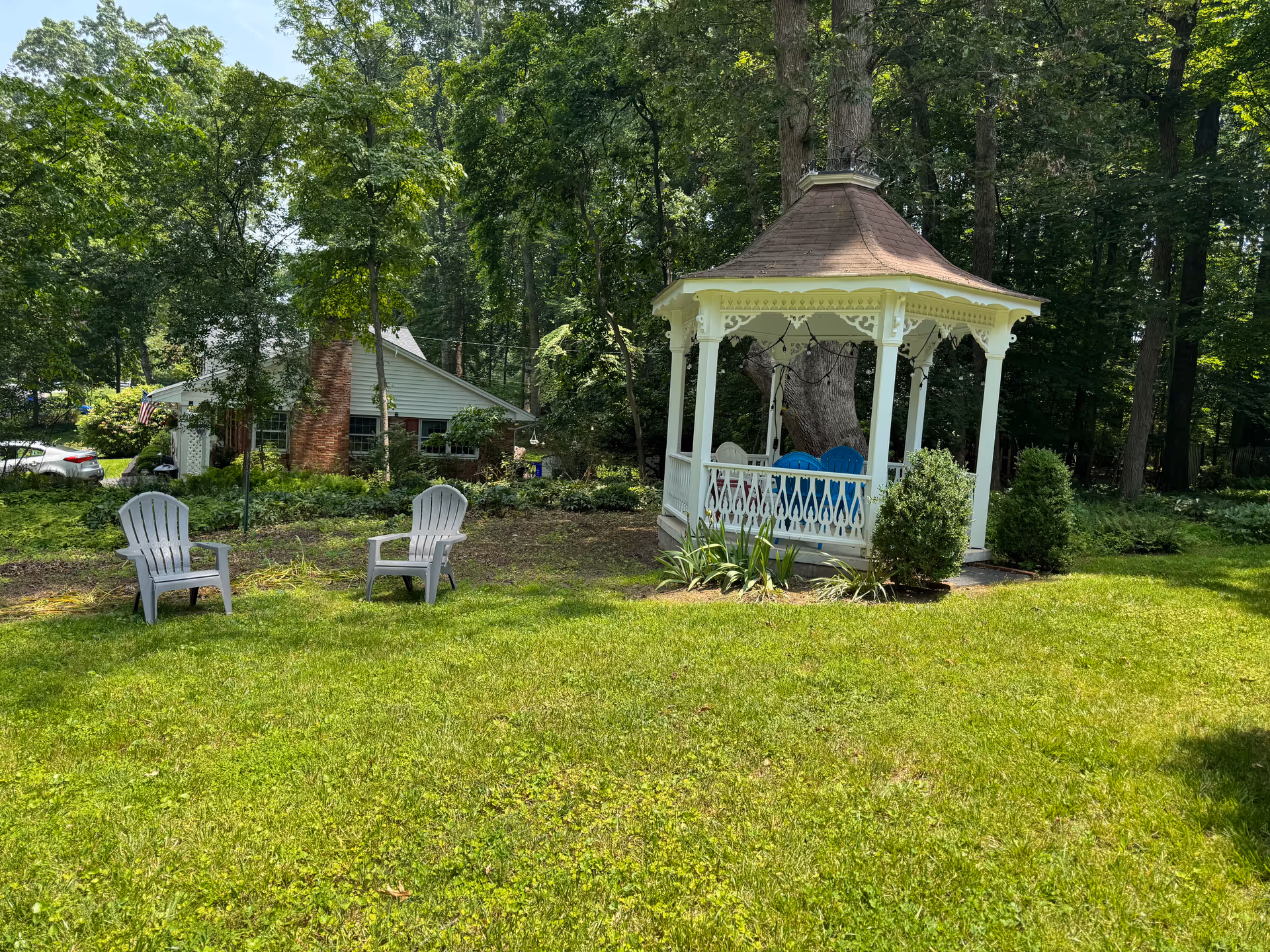 Sunlit yard with a white gazebo, two chairs, a house in the background, and trees.