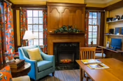 Cozy living room with a lit fireplace, a blue armchair, wooden mantel and built-in desk beside windows.