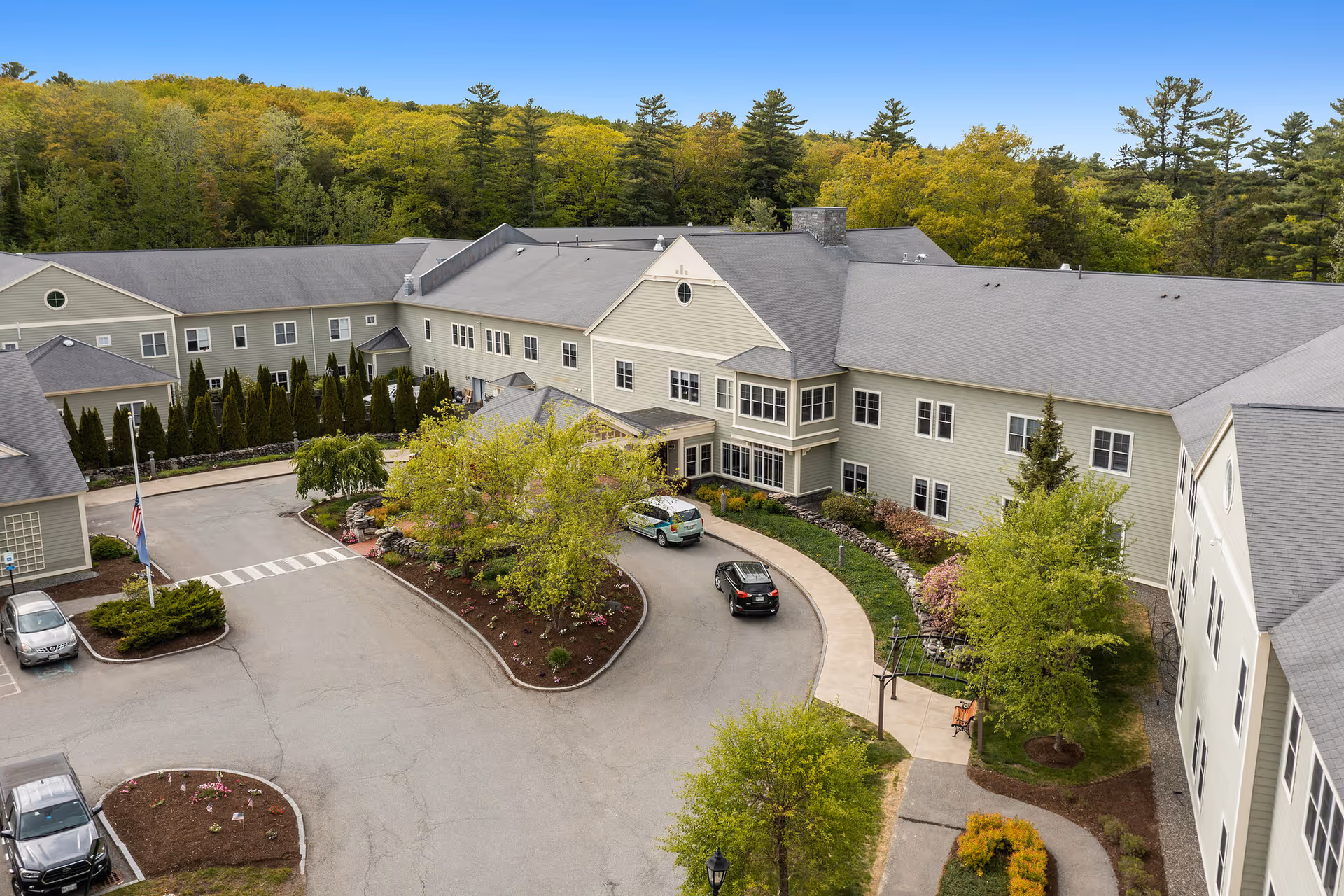 Aerial view of Dirigo Pines senior living facility showing a large, two-story building with gray siding and a gray roof surrounded by trees and greenery. There is a circular driveway with a few parked cars and landscaped areas with bushes and small trees.