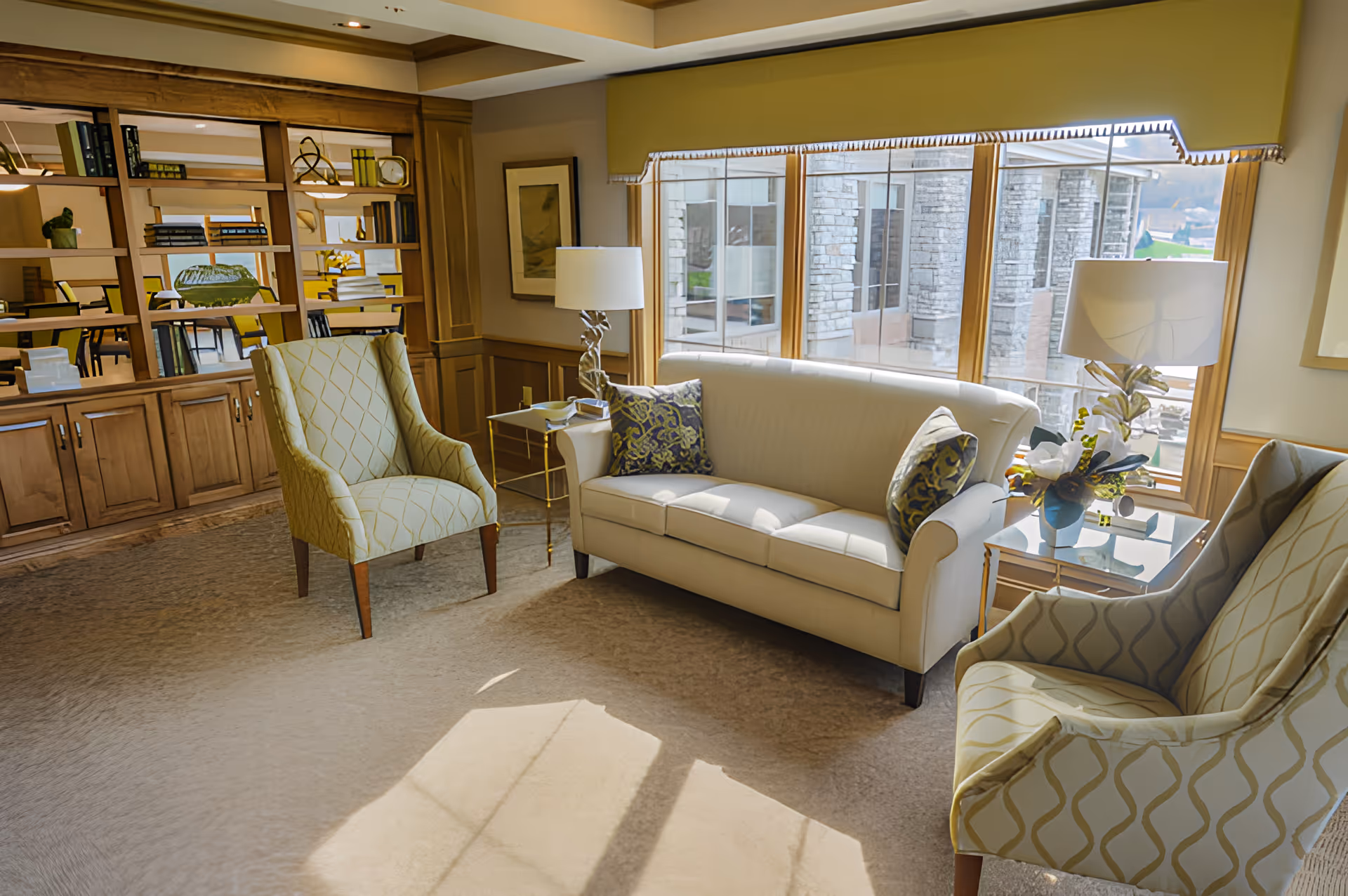 Sunlit communal living room with a cream sofa, patterned armchairs, side tables and large windows looking out to a stone-clad exterior.