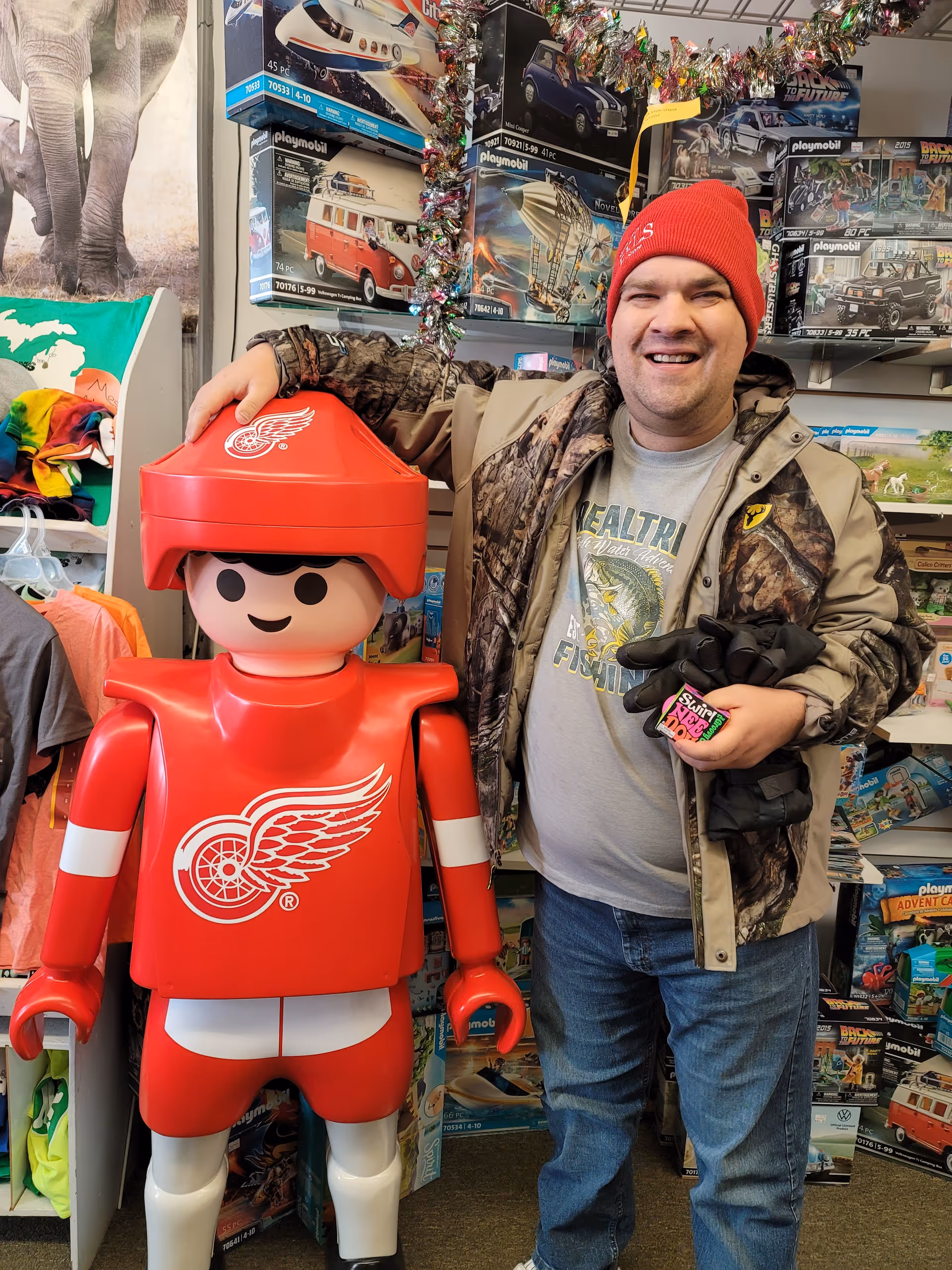 Man in camo jacket and red beanie stands beside a large red toy robot.