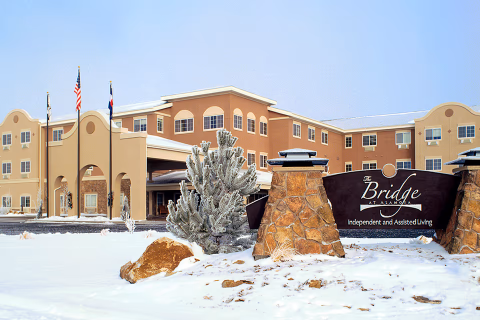 Snow-covered front of The Bridge at Alamosa senior living facility with flags, an entrance canopy, and a stone sign.