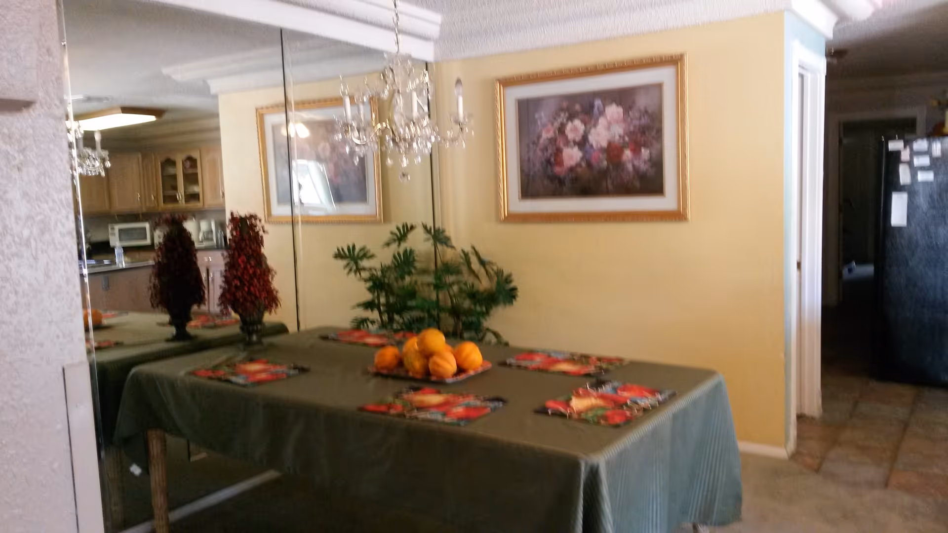 Dining room with a green-clothed table laid with colorful placemats and a bowl of fruit, a mirrored wall and floral painting, and the kitchen visible in the background.
