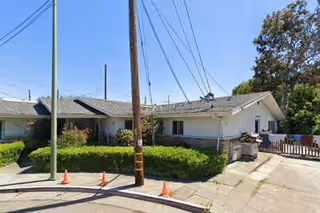 Exterior view of a single-story residential building with a sloped roof, surrounded by greenery and a paved driveway. There are utility poles and power lines visible in front of the building.