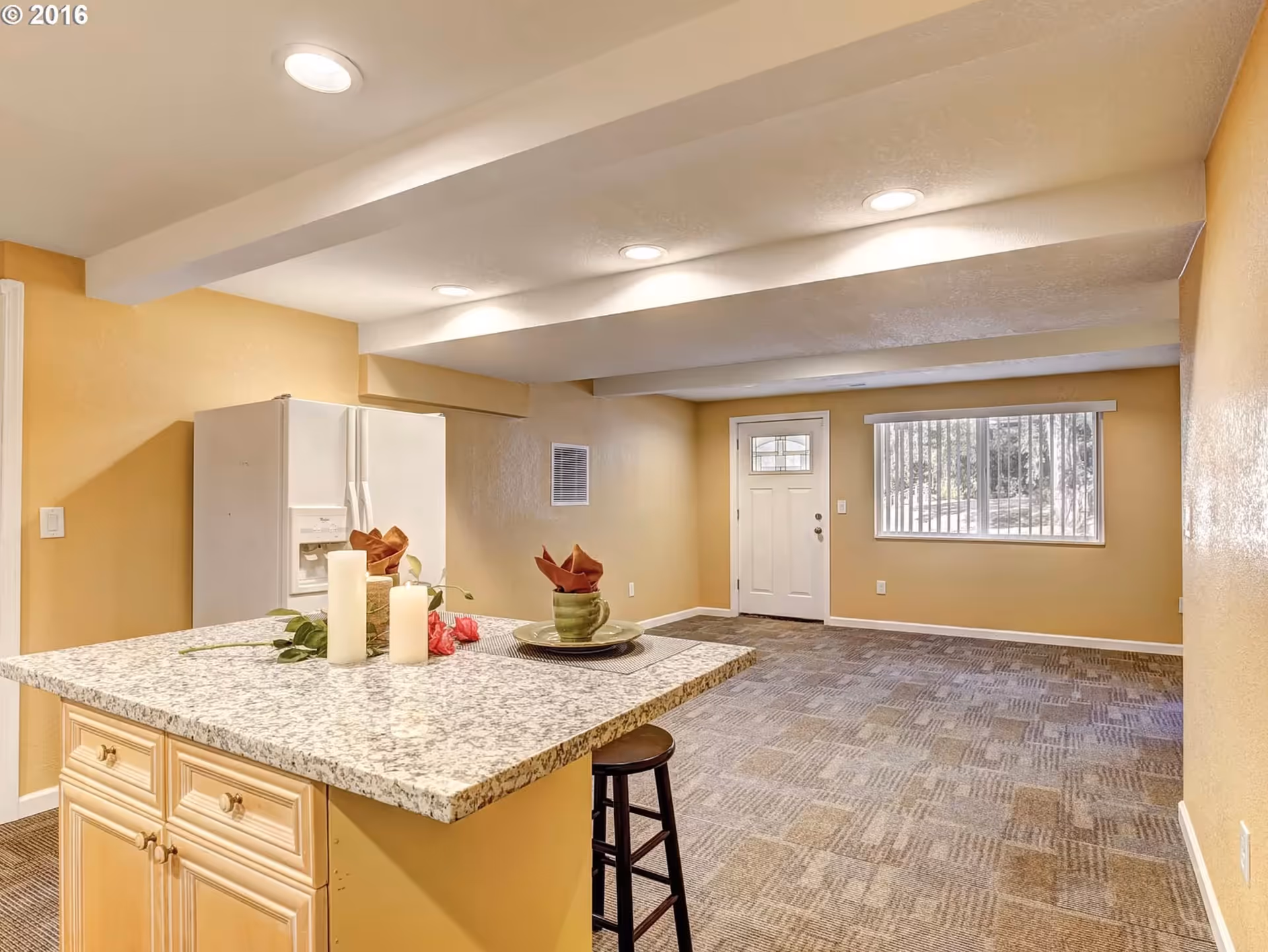 Bright kitchen area with granite island, refrigerator, and doorway in warm beige room.
