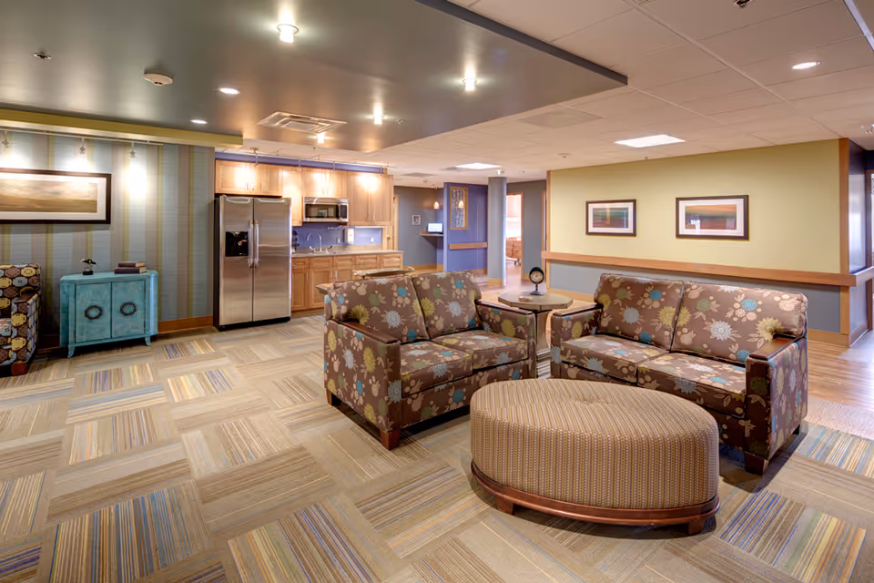 Bright senior living lounge with patterned sofas, ottoman, and kitchenette in the background.