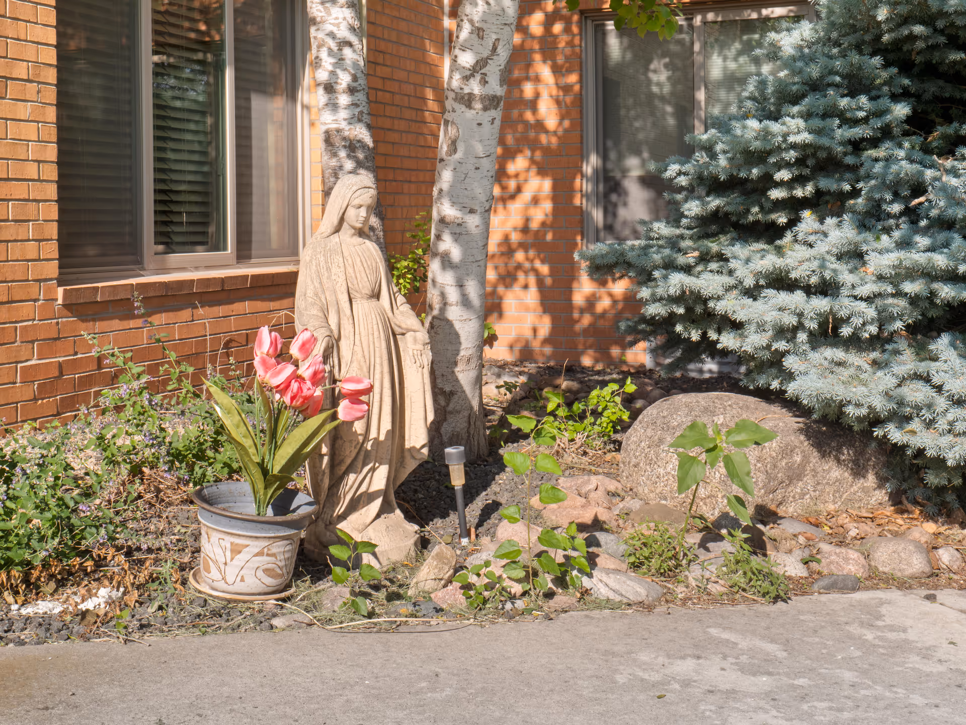 Exterior brick wall with window, statue, potted tulips, and a small garden area.