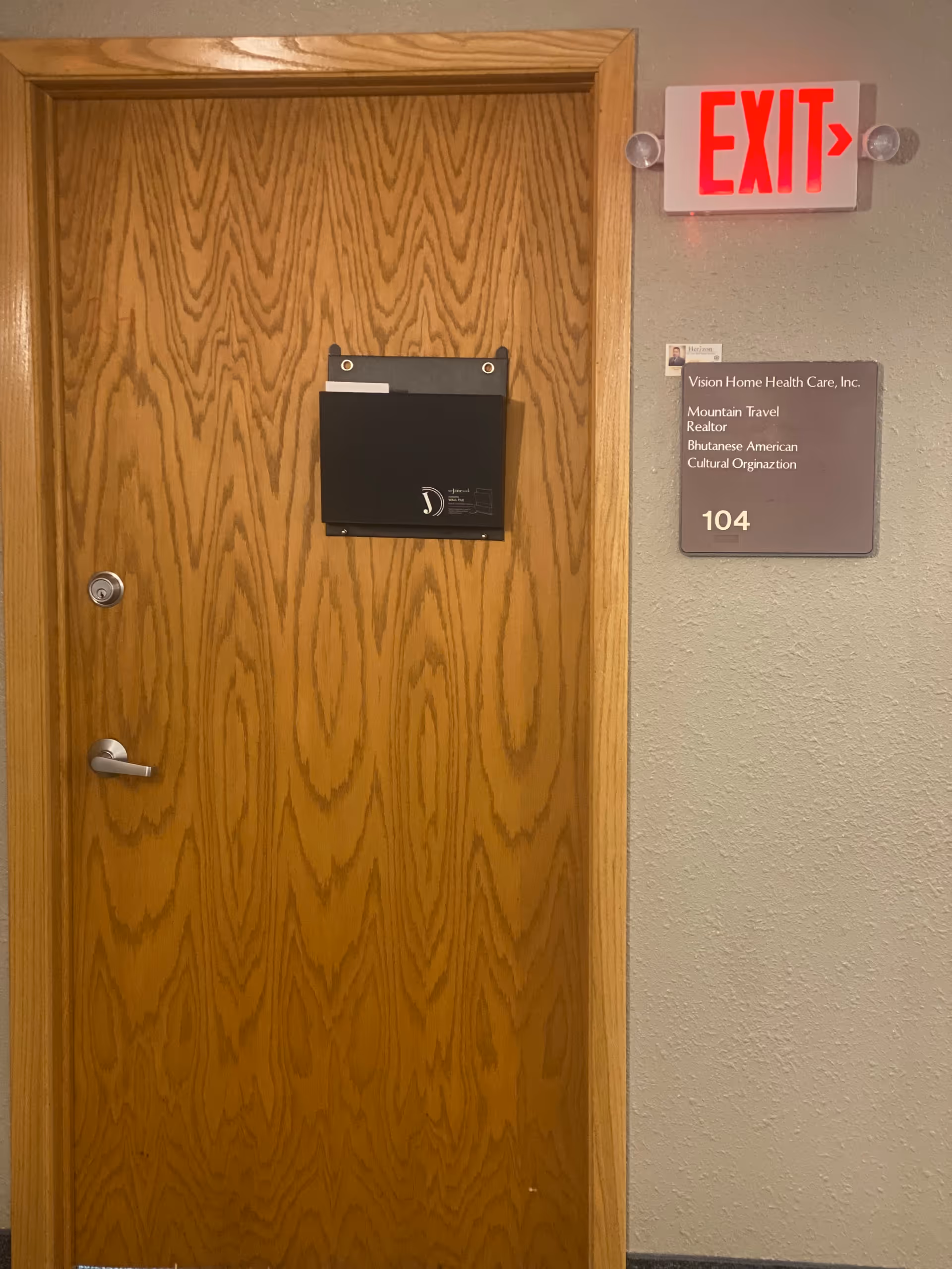 Wooden door in hallway with black wall mail holder, exit sign, and Vision Home Health plaque.