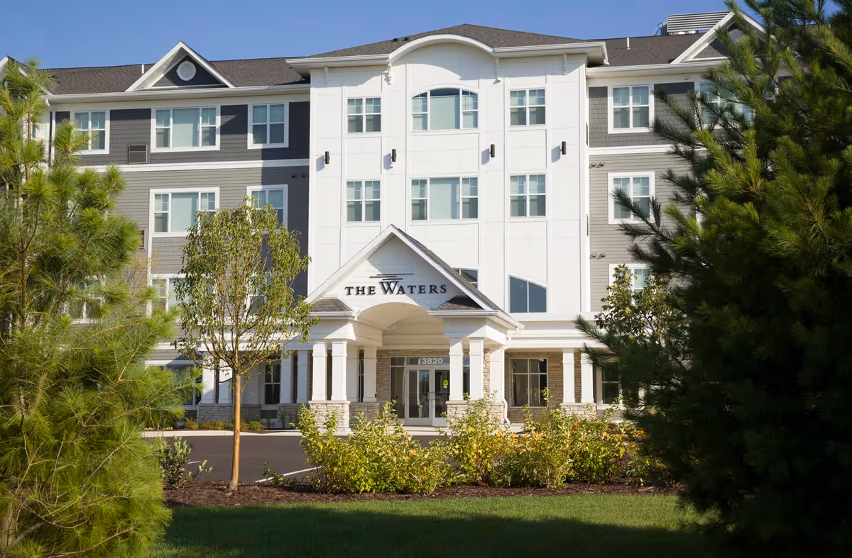 Front exterior view of a senior living facility named The Waters, a multi-story building with white and gray siding, surrounded by trees and landscaping under a clear blue sky.