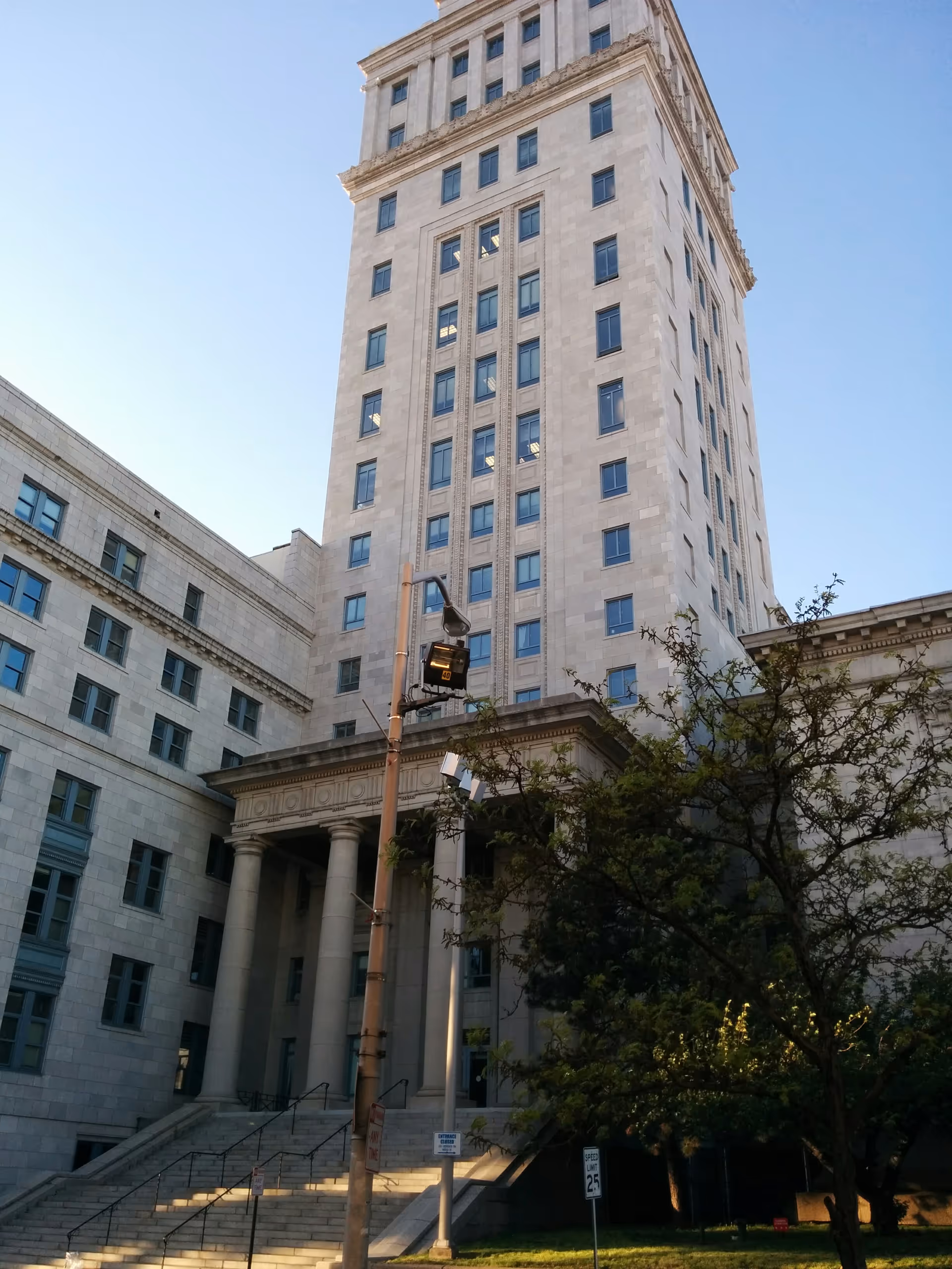 Tall stone building with classical columns, stairs, and a tree under a clear sky.