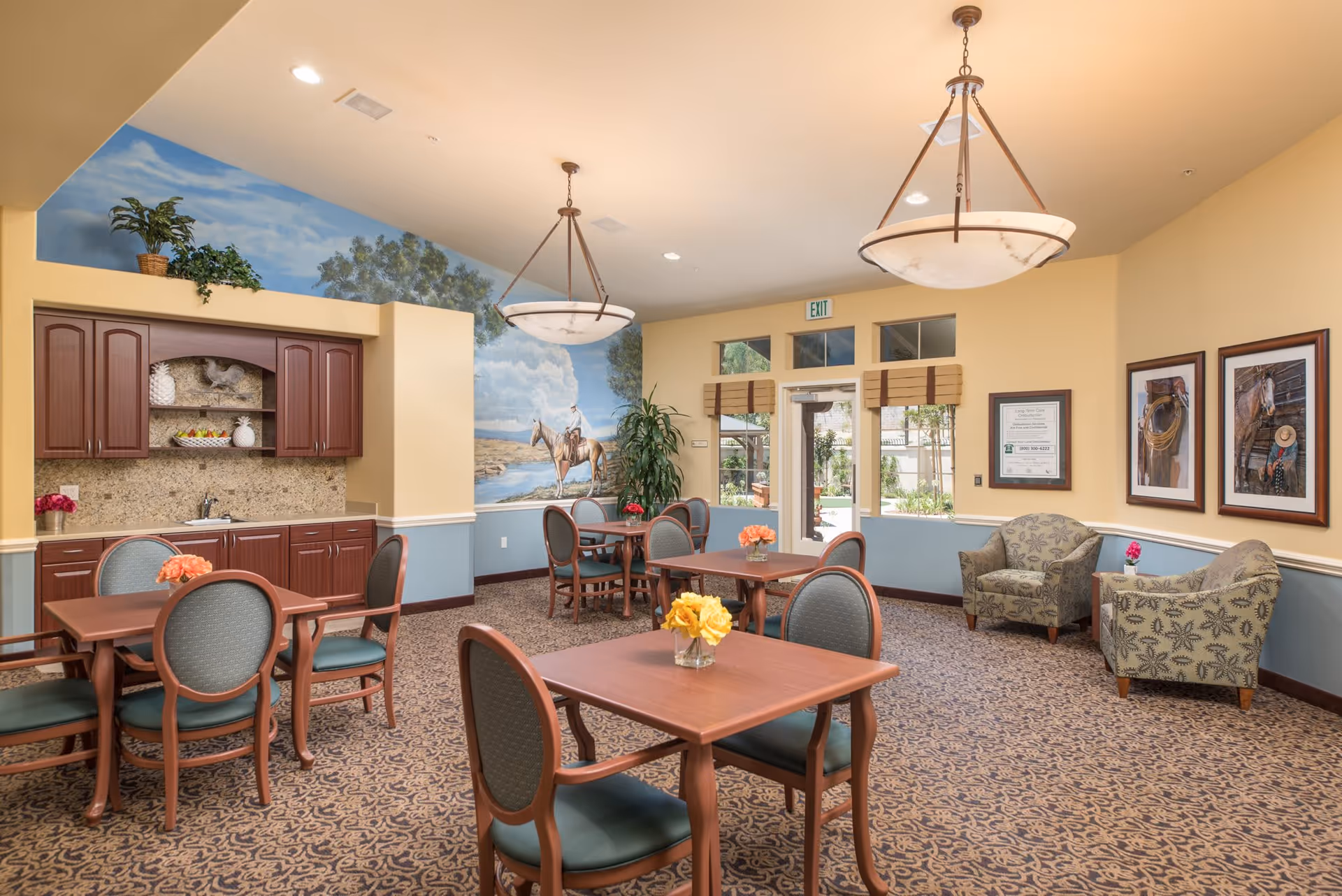 A bright and inviting common area with several wooden tables and chairs arranged on a patterned carpet. The walls are painted yellow and blue with a mural of a person on horseback near a river. There are two hanging light fixtures on the ceiling, framed pictures on the walls, and a small kitchenette with cabinets and a sink. A glass door and windows provide a view of an outdoor garden area. Two upholstered armchairs and a small table with a flower vase are placed in one corner.