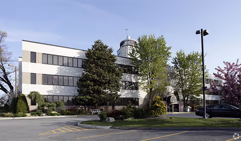 Two-story senior living facility with a white and gray facade, trees, and a parking lot.