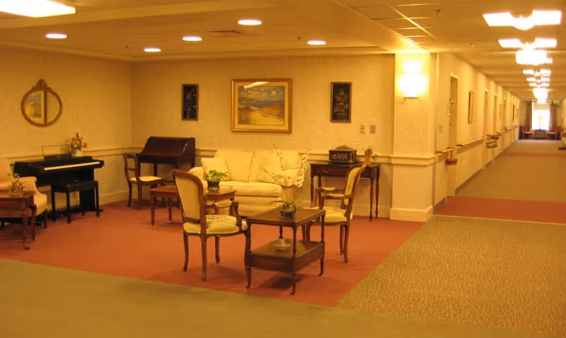 Warmly lit senior living common area with a sofa, chairs, small tables, a piano, and a long hallway.