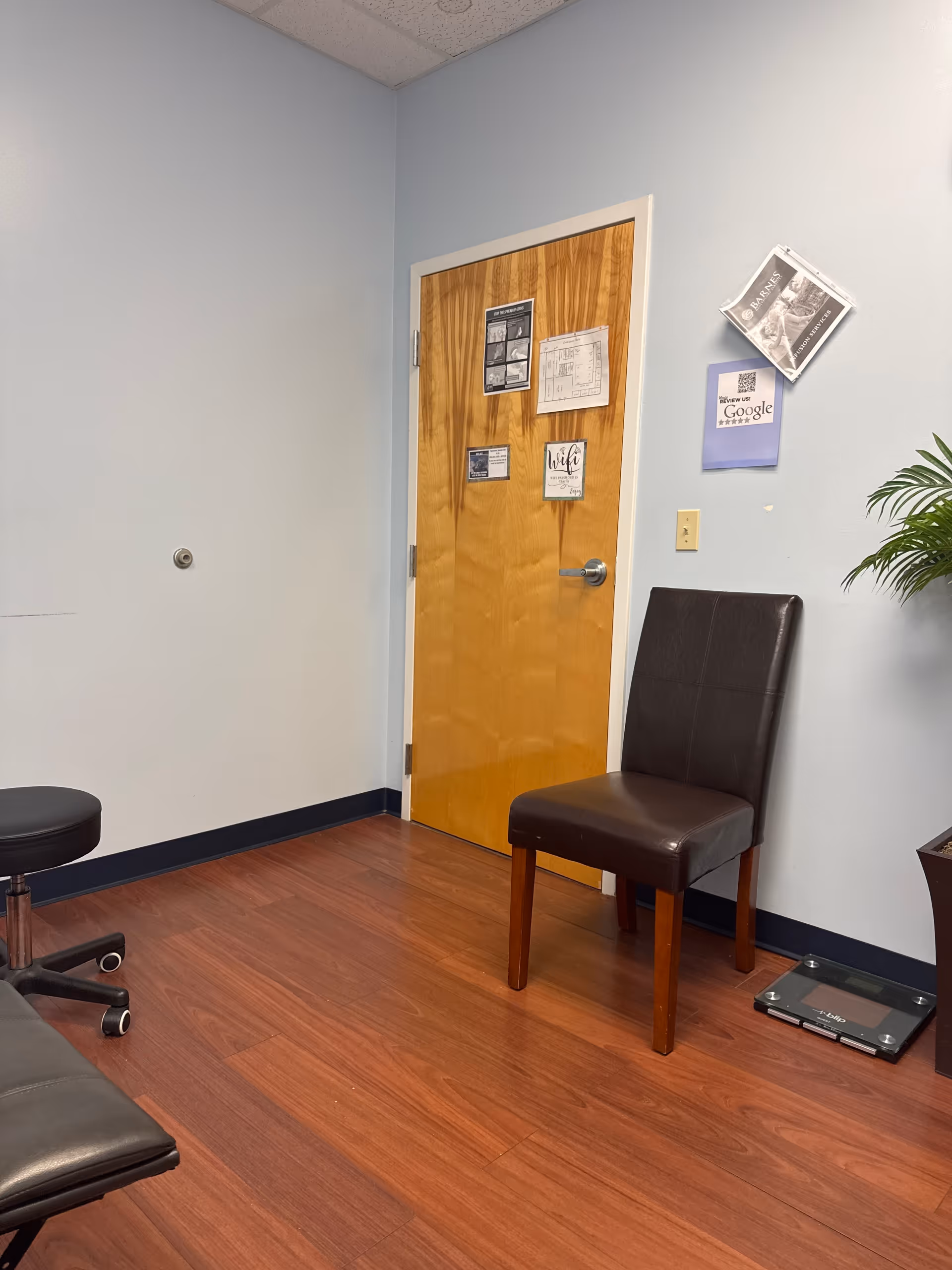 A small interior room with light blue walls, a wooden door, chair, scale, and plant.