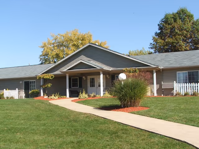 Single-story residential building with a covered central entrance, concrete walkway, and manicured lawn under a clear blue sky.