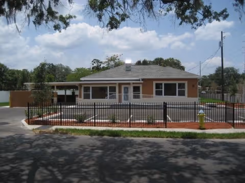 Single-story building with a gray roof and light brown exterior, surrounded by a black metal fence. There is a parking area in front with small landscaped areas containing plants and mulch. A yellow fire hydrant is visible near the fence, and trees and power lines are in the background under a partly cloudy sky.