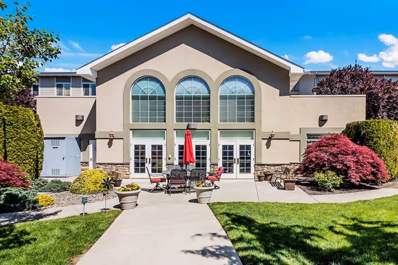 Exterior view of a building with three large arched windows above three sets of French doors. In front of the building, there is a concrete pathway leading to an outdoor seating area with a table, chairs, and a red umbrella. The surrounding area features well-maintained landscaping with green grass, bushes, and small trees under a clear blue sky.