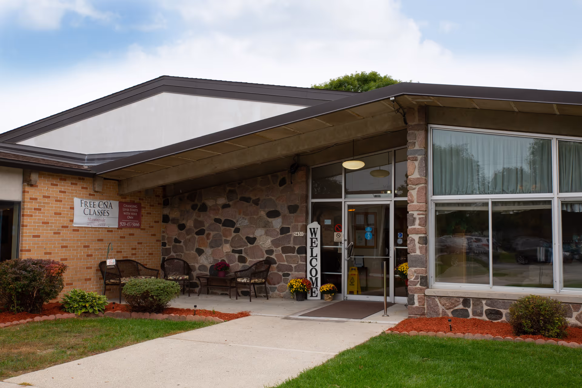 Exterior view of a building entrance with stone and brick walls, a glass door, and large windows. There is a vertical 'WELCOME' sign next to the door, outdoor seating with chairs and a table, and some potted plants and landscaping with grass and mulch. A sign on the brick wall advertises free CNA classes.